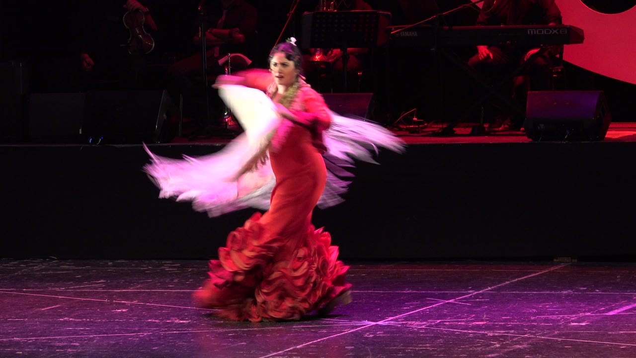 Woman in a red ruffled dress swinging a white shawl on a dark stage.