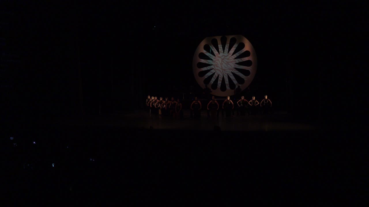 Opening frame of a Cabo San Lucas stage performance, with small performers arranged beneath a dimly lit circular wheel prop and a nearly black audience foreground.