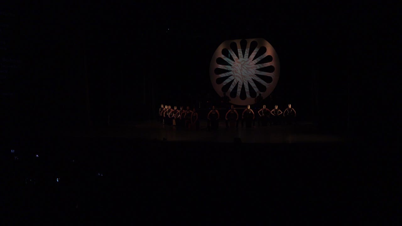 Cabo San Lucas performance frame with the ensemble still grouped across the lower stage and the circular wheel prop faintly lit above them.