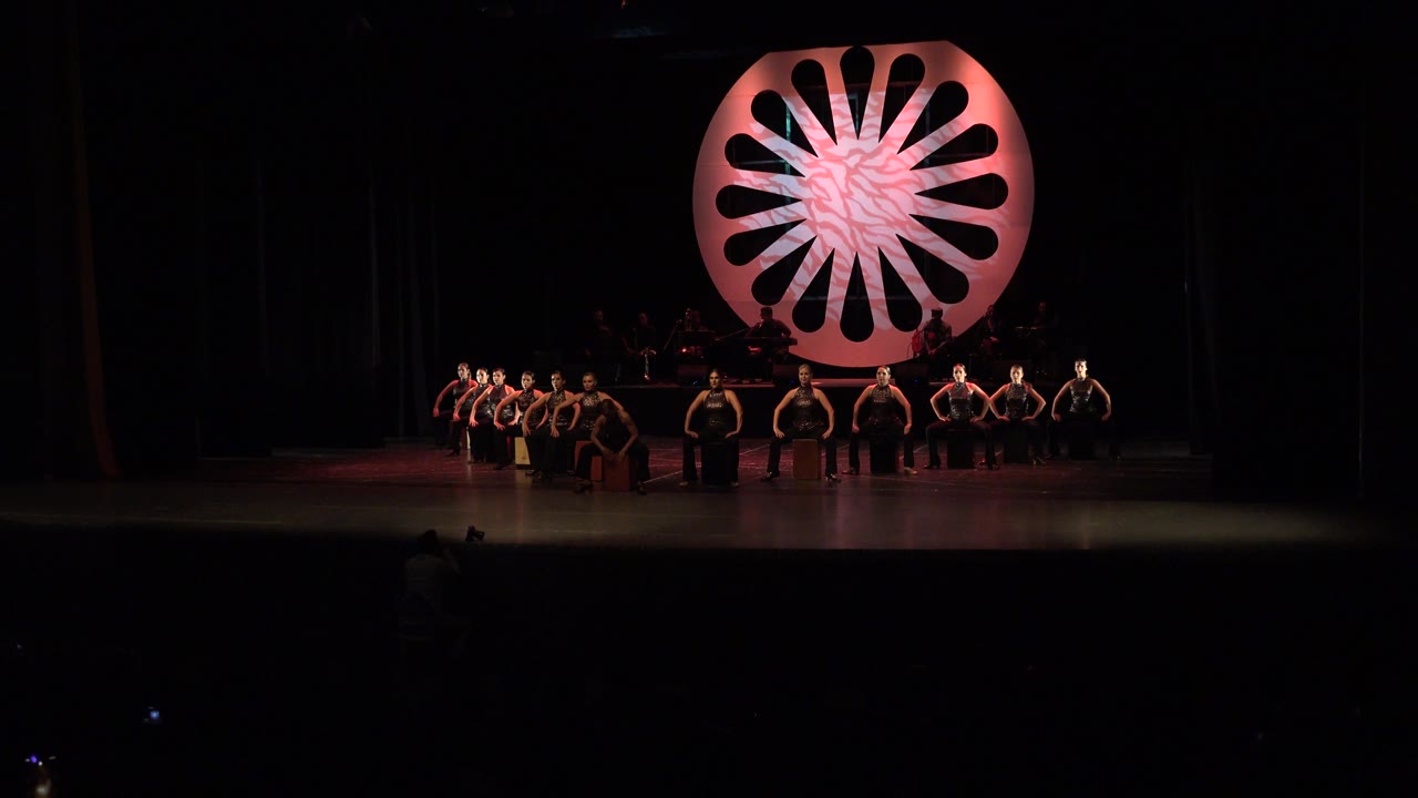 Cabo San Lucas performance frame with the ensemble still spread across the stage, the band dimly visible behind them, and the pink circular set piece glowing overhead.