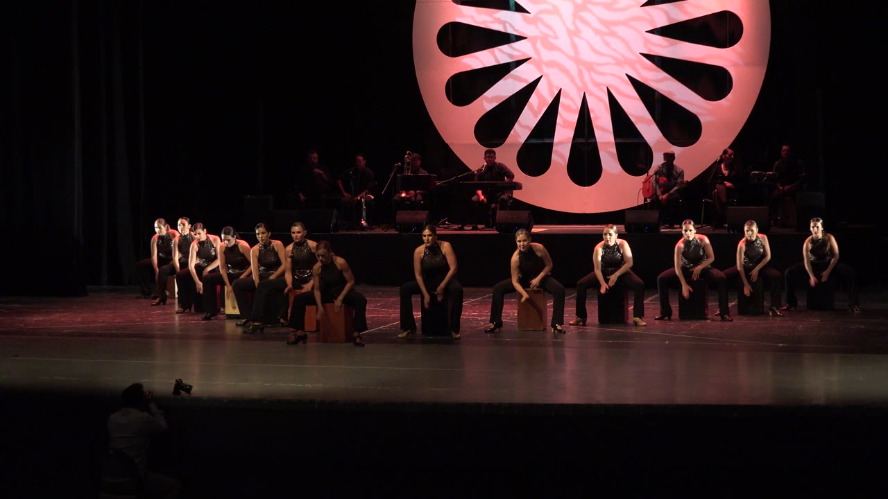 Wide stage view with women performers seated under a pink circular backdrop.