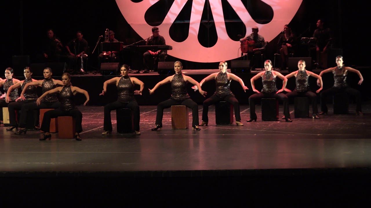 Women in matching black outfits perform seated choreography on a Cabo San Lucas stage, with musicians in shadow and a large pink backdrop behind them.