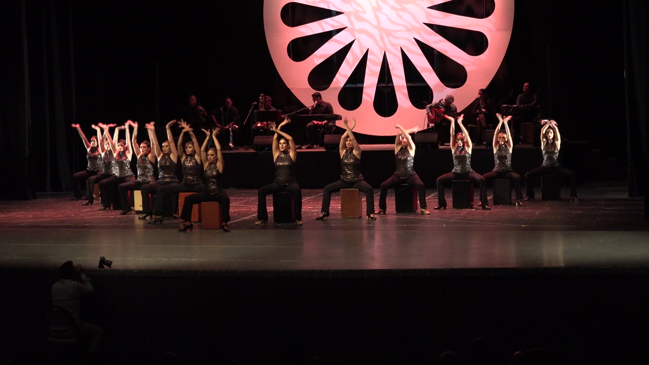In Cabo San Lucas, Mexico, a wide stage view shows seated dancers lifting their arms while musicians sit in shadow behind a large red-pink circular backdrop.