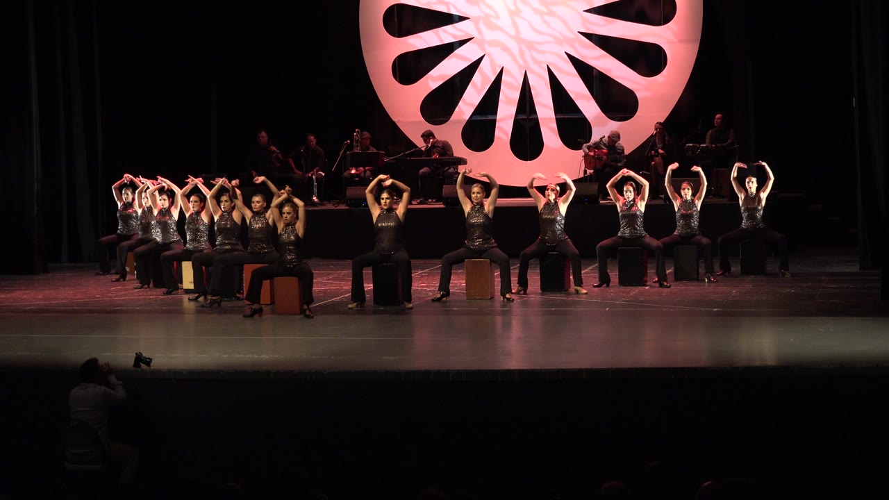 Dancers form overhead arm shapes while seated across the stage.