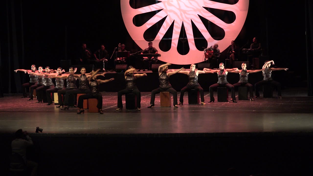 Dancers in black costumes sit across a stage with arms extended.