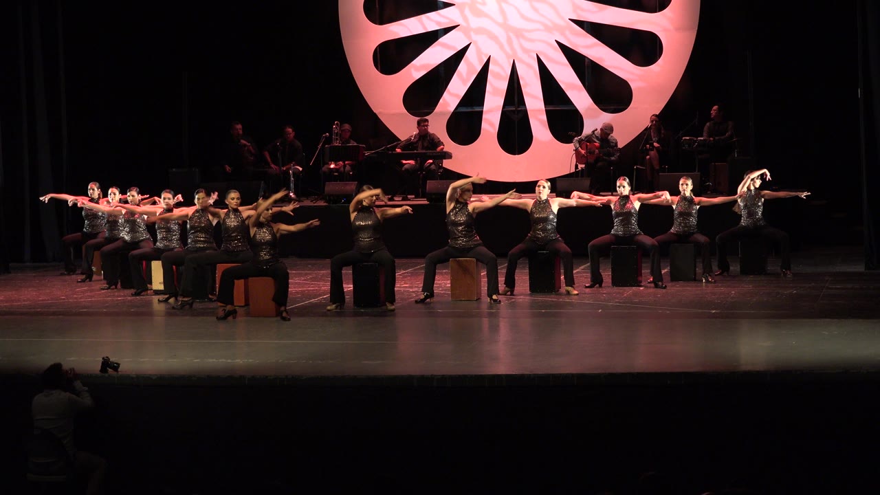 Wide Cabo San Lucas stage frame with the seated dancers maintaining long horizontal arm lines while the musicians remain dimly lit behind them.