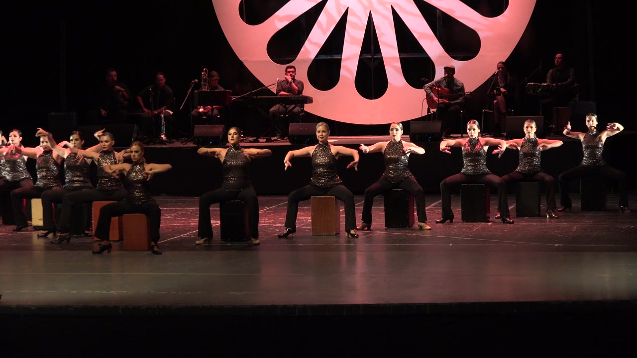 Wide view of adult women in black stage outfits seated across a Cabo San Lucas theater stage, with musicians in shadow and a large pink circular backdrop above them.