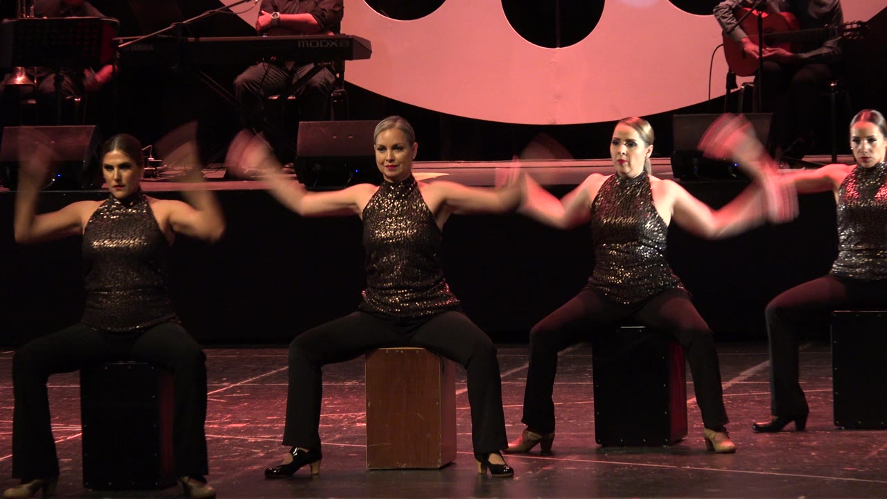 Adult women in black performance outfits extend their arms while seated on stools on a Cabo San Lucas theater stage with red light across the backdrop.