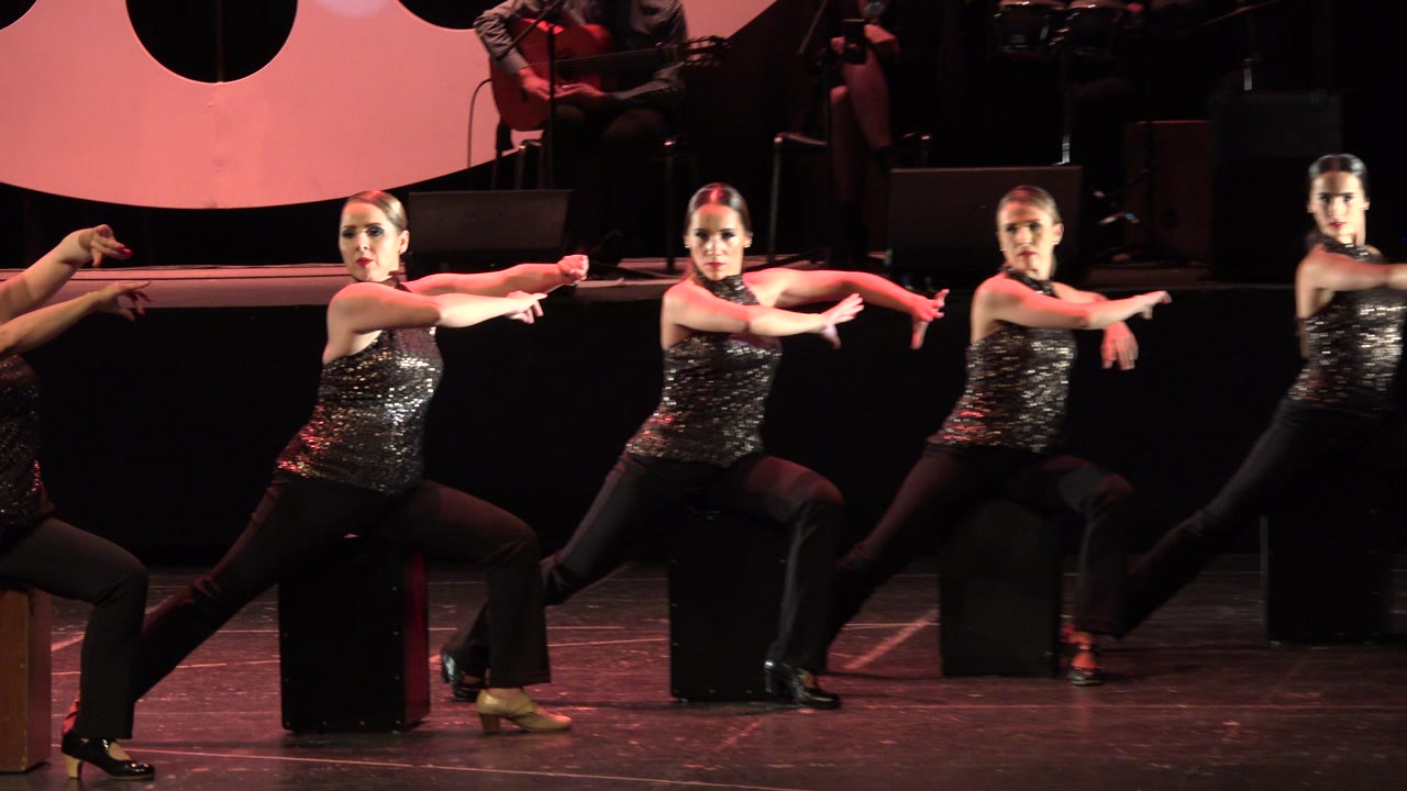 Women in black stage outfits perform seated dance choreography.