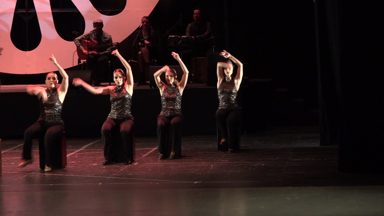 Four seated female dancers on a stage with musicians behind them.