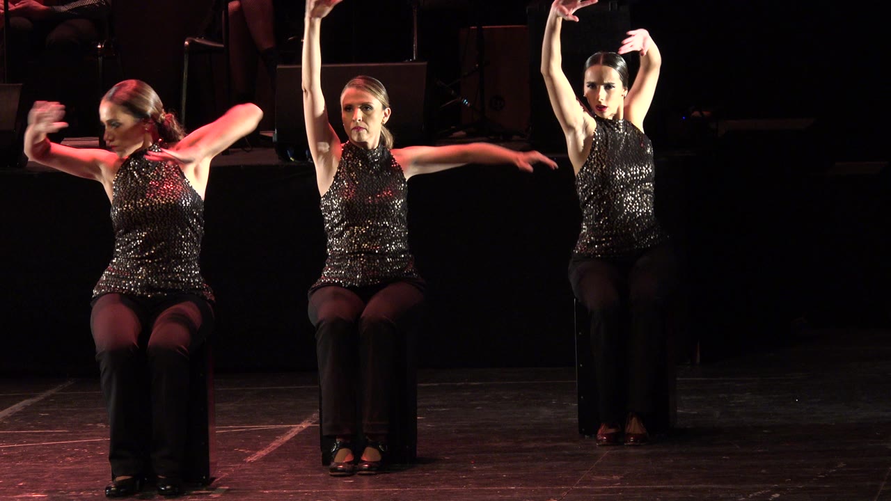 Three women perform seated choreography on a dark stage in Cabo San Lucas, Mexico, with sequined black tops and warm red side lighting.