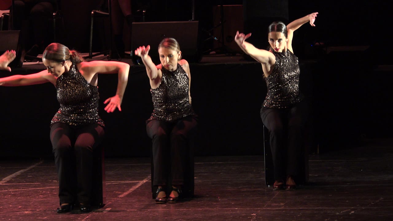 Three seated women gesture forward and sideways on a dark stage.