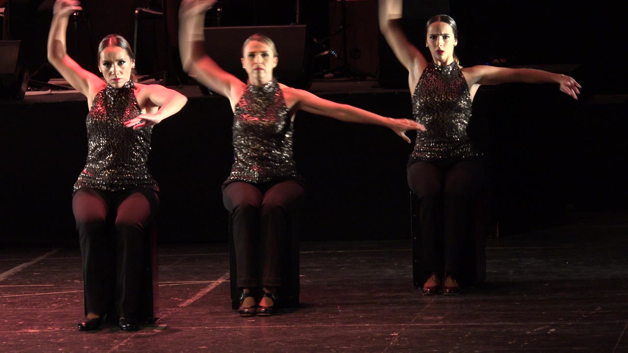 Three seated women performing arm choreography on a dark stage.