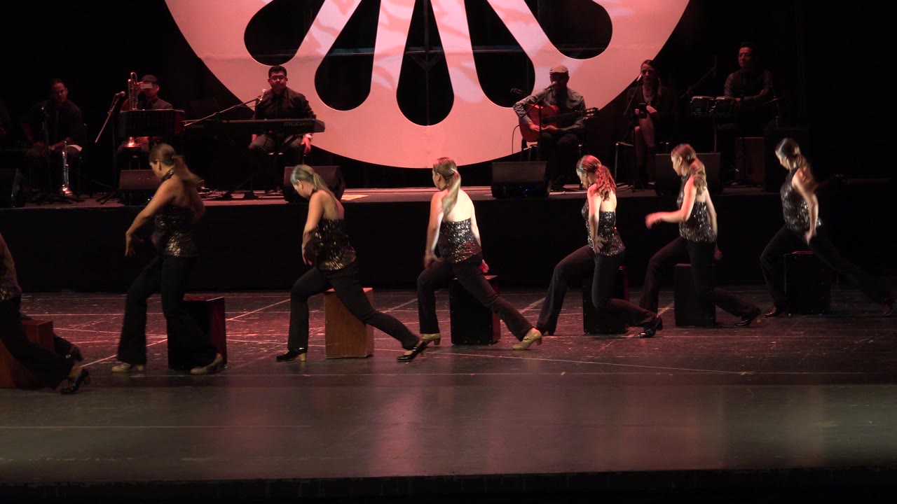 Women crossing a stage in front of seated musicians and a red circular backdrop.