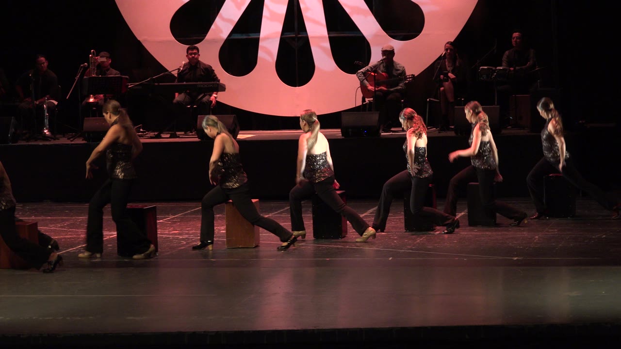 Several women continue moving across the Cabo San Lucas stage near small box seats, with silhouetted orchestra members and the red backdrop behind them.
