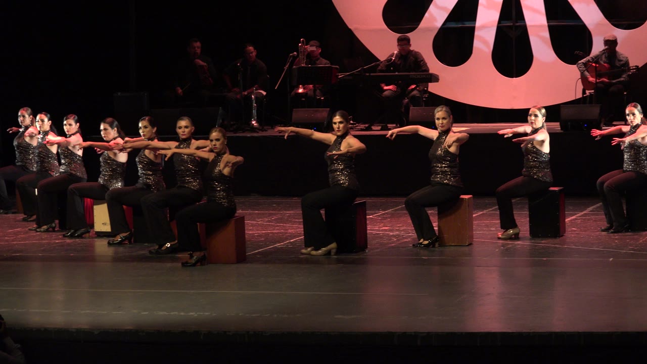 At the start of the Cabo San Lucas performance, seated dancers in dark sparkling tops extend their arms in a line while musicians are visible in the background.