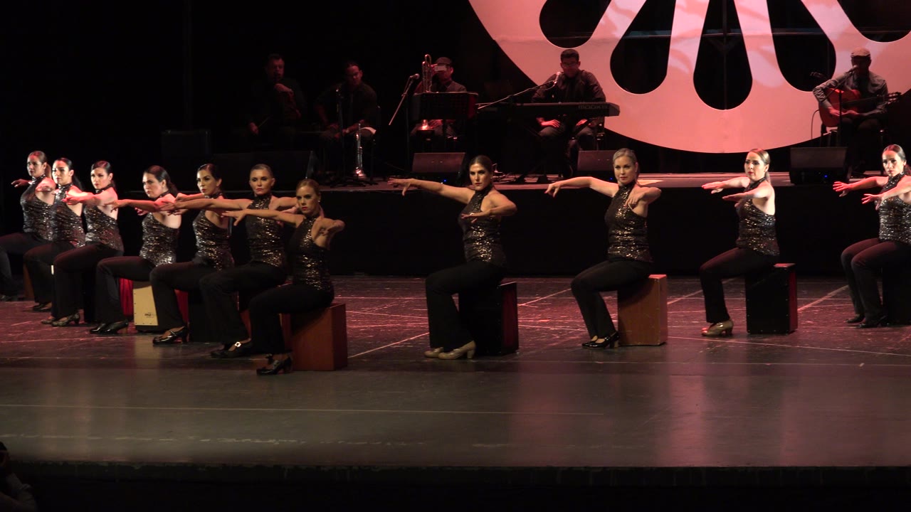 One second in, the Cabo San Lucas dance group holds a wide seated formation with arms stretched outward and the live band dimly lit behind them.
