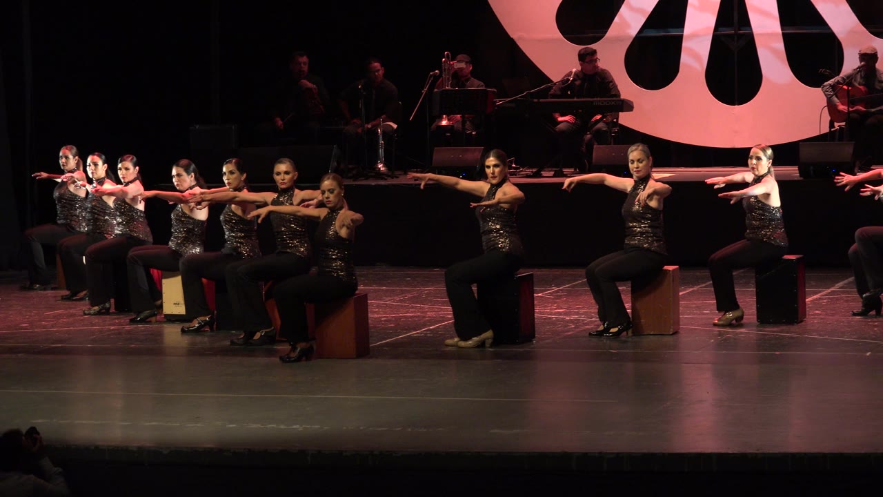 A row of seated dancers points outward under red stage lighting.