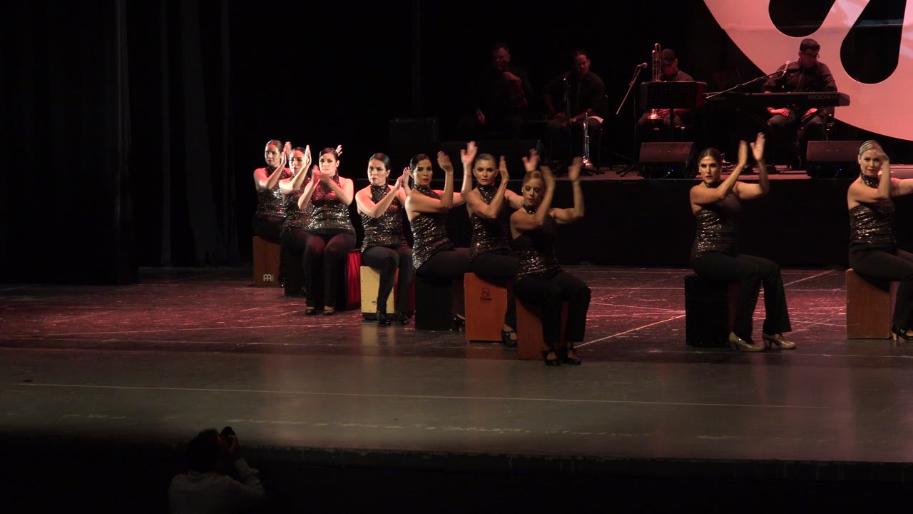 Women seated on box drums perform synchronized hand movements on a dark stage.