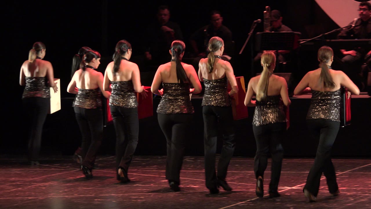Women dancers walk away from camera across a stage carrying square props.