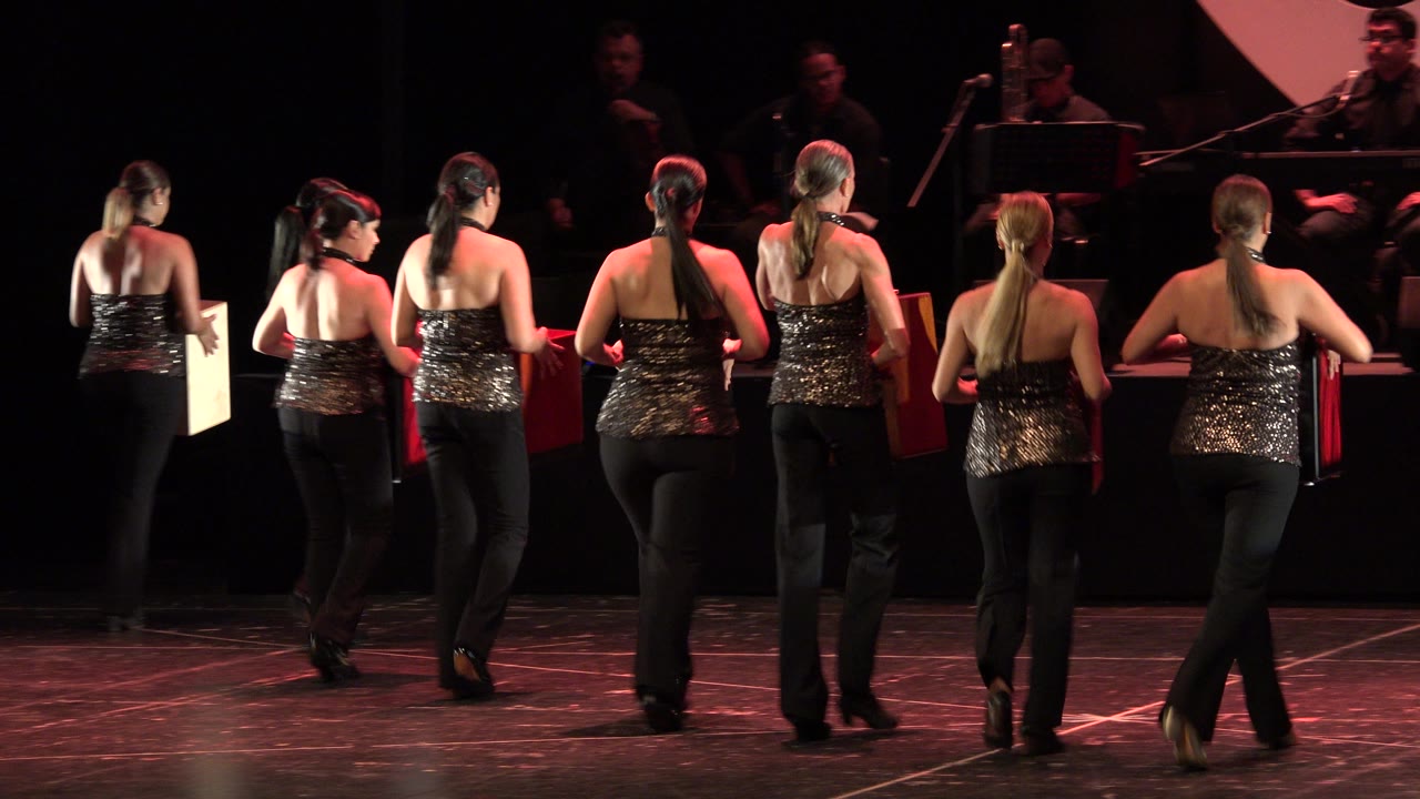 The group continues moving across the Cabo San Lucas stage, with several dancers seen from behind and a band positioned in the darker background.