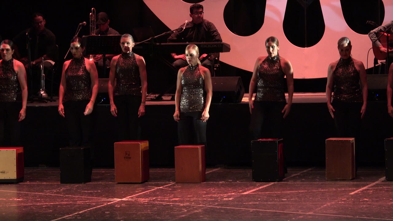 Adult women in matching black sleeveless tops and black pants stand across a stage in Cabo San Lucas, Mexico, with musicians, microphones, and box percussion stools visible behind and in front.