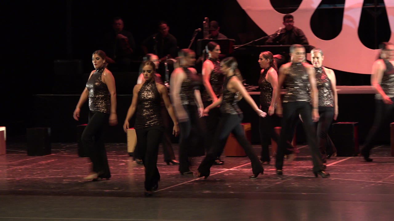 A wide stage view shows women continuing their dance entry in Cabo San Lucas, Mexico, while background musicians sit in low light.