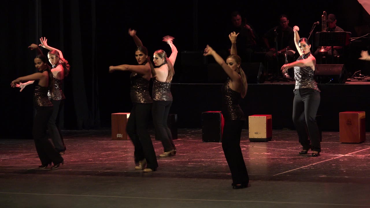 Women in matching dark outfits dance on a stage with musicians behind them.
