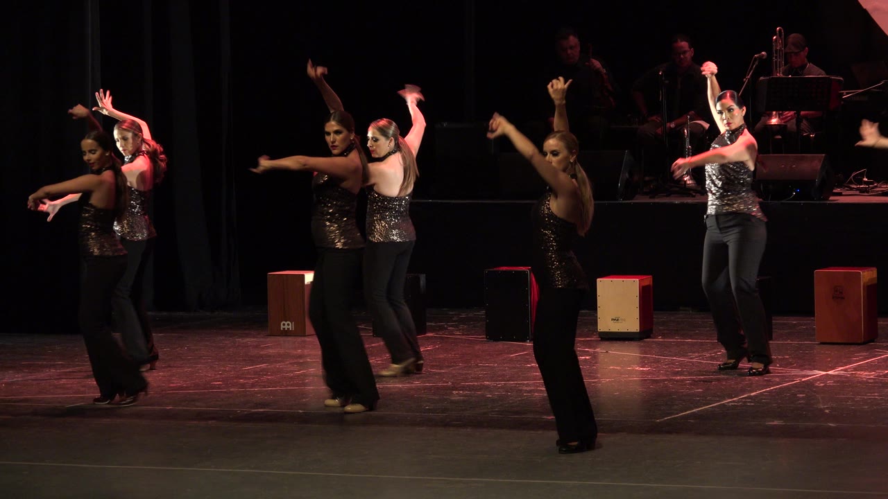 The dance group continues the routine in Cabo San Lucas, with several women angled toward the audience and one dancer at right framed near the musicians.