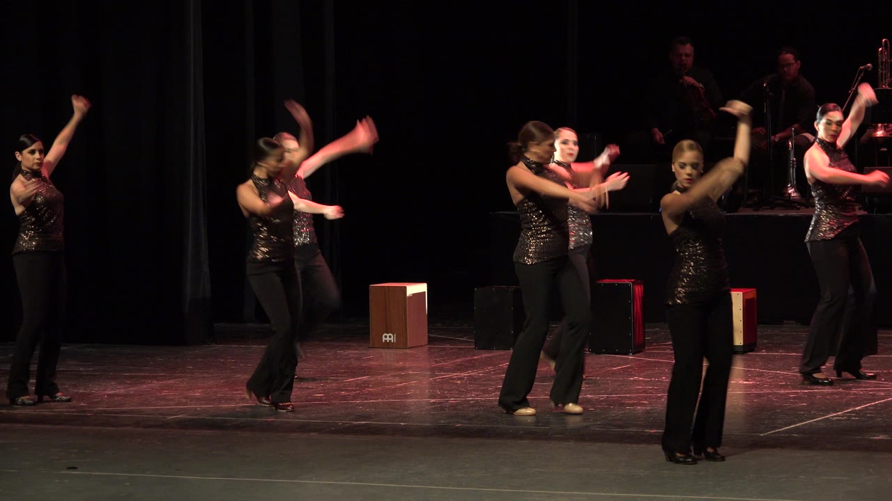 Several women continue the choreographed routine on the theater stage in Cabo San Lucas, Mexico, with arms extended and percussion equipment visible near the rear curtain.