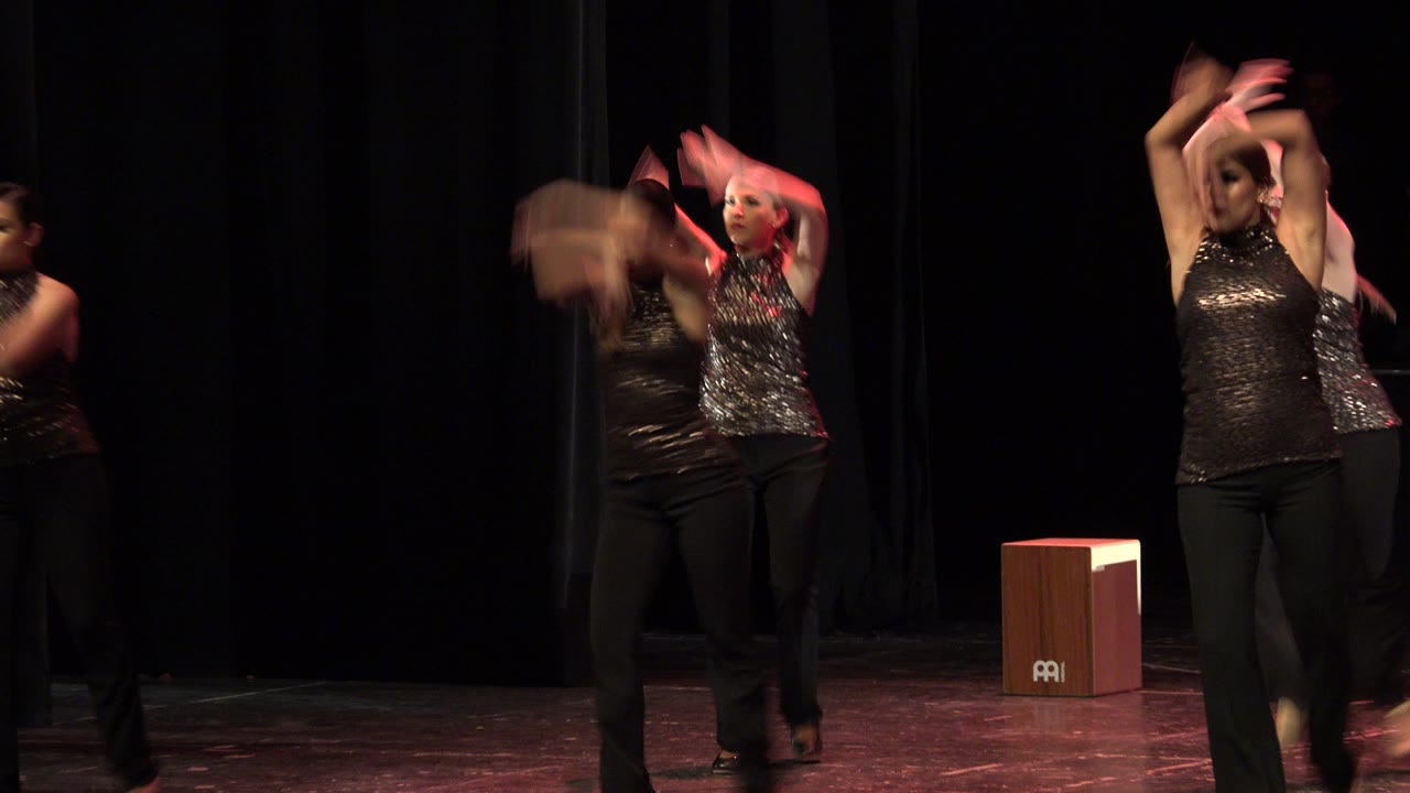 Two adult women in black performance outfits lift their arms on a Cabo San Lucas stage, with a wooden cajon placed behind them near the right side of frame.