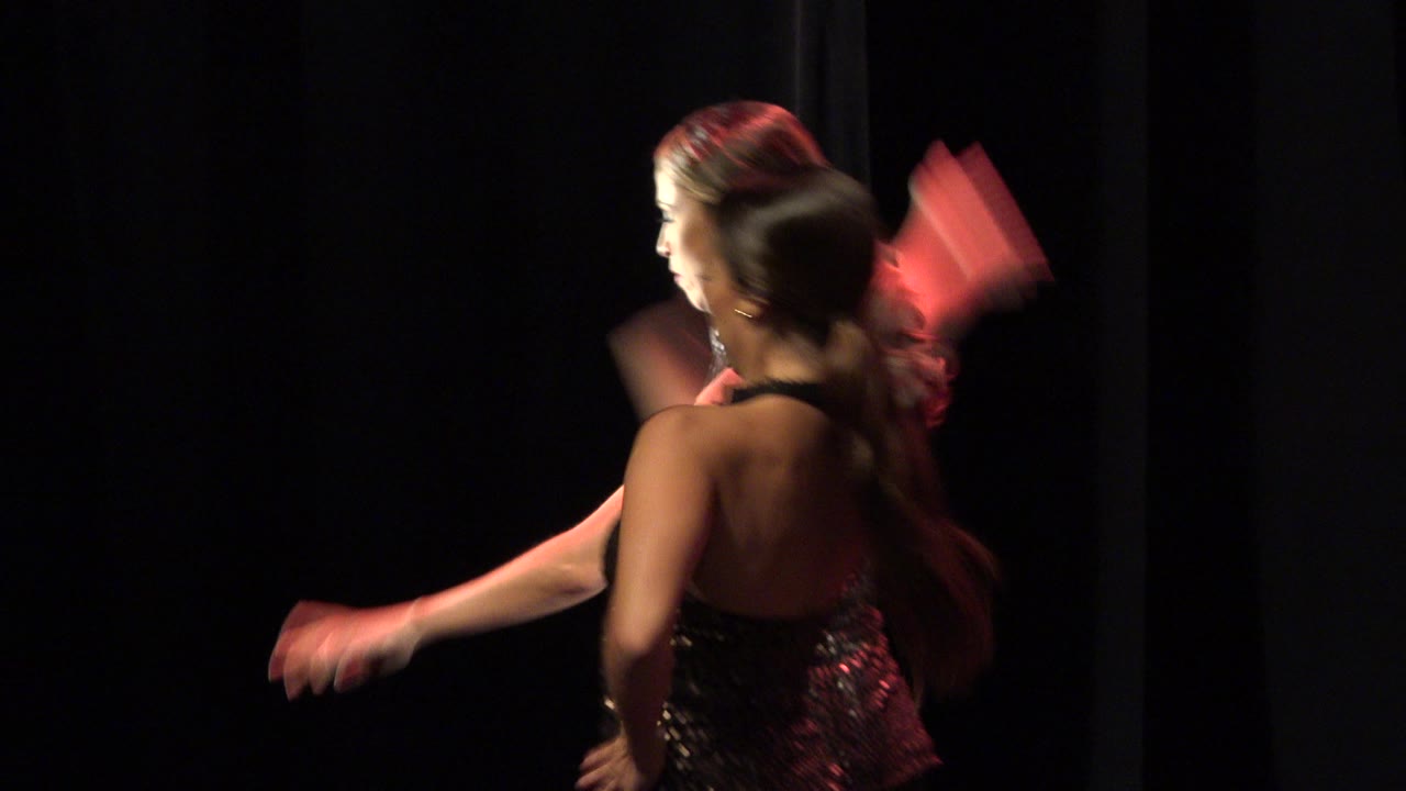 Two women continue the choreographed dance in Cabo San Lucas, Mexico, with blurred arms, black curtains, and red light catching the gold costume details.