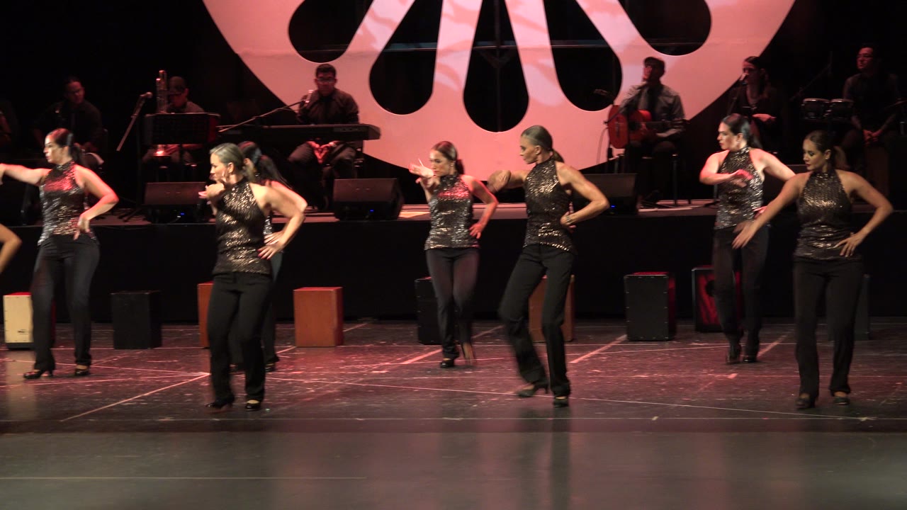 Female dancers perform on a stage with musicians behind them.