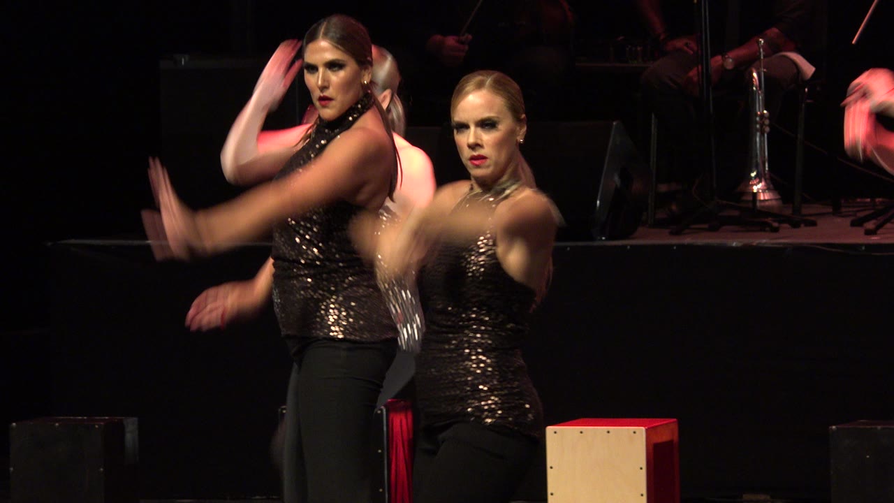 Two women in glittering tops dance on a dark stage.