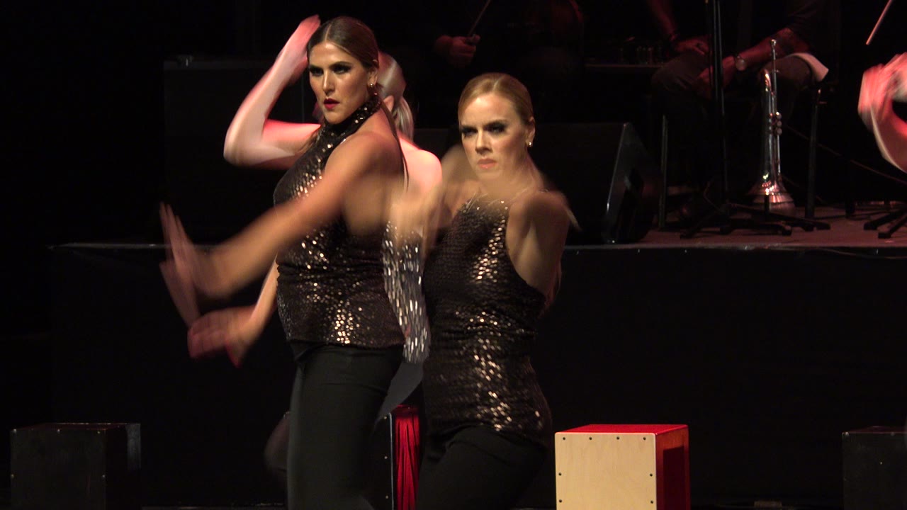 Two adult women in matching dark performance outfits face the camera during the Cabo San Lucas stage routine, with a light wood percussion box at the rear edge of frame.