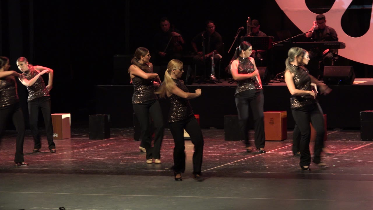 Adult women continue a synchronized dance phrase on the Cabo San Lucas stage while a large pink-and-black backdrop shape frames the band area.
