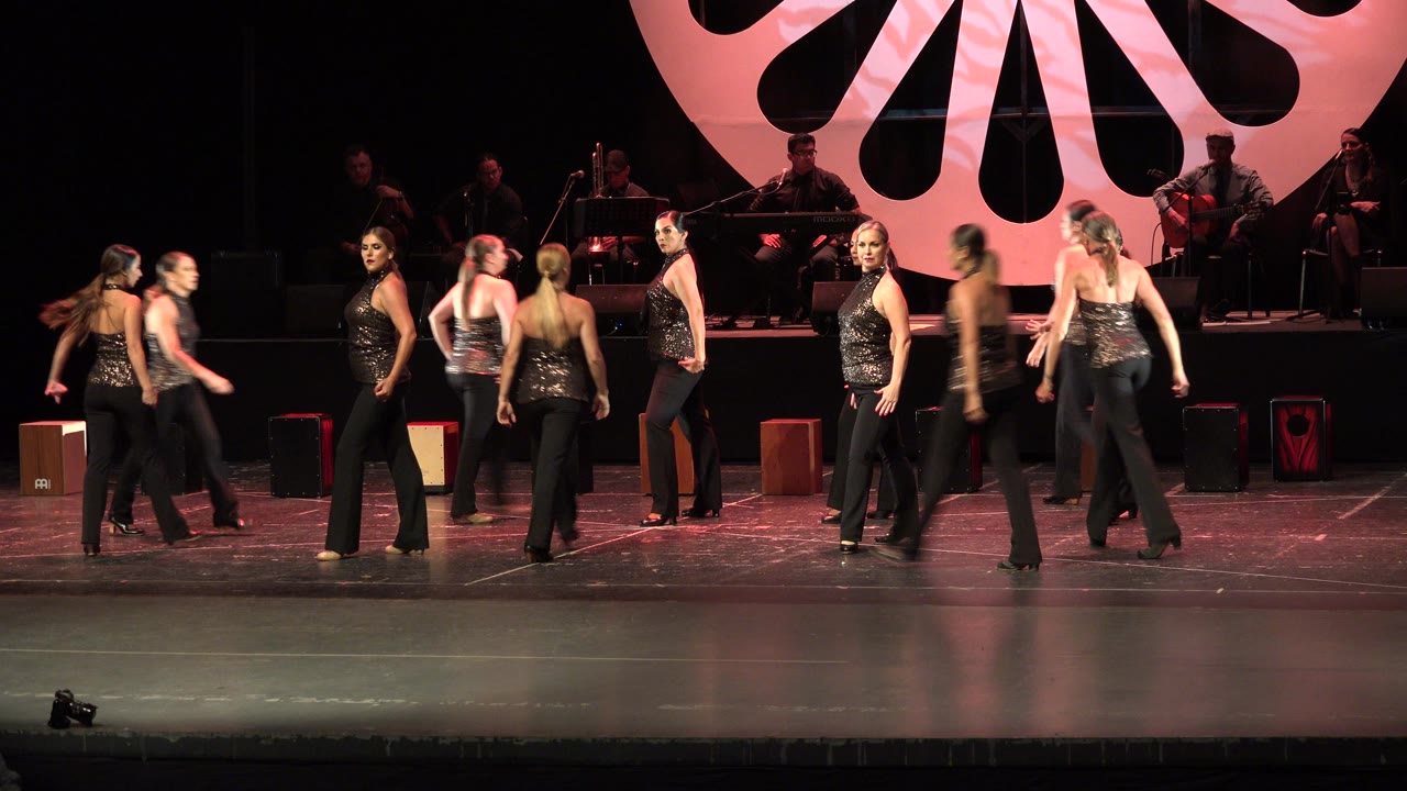 At 0:00 in Cabo San Lucas, dancers in black outfits move across a pink-lit stage while musicians sit behind a large fan-shaped backdrop.