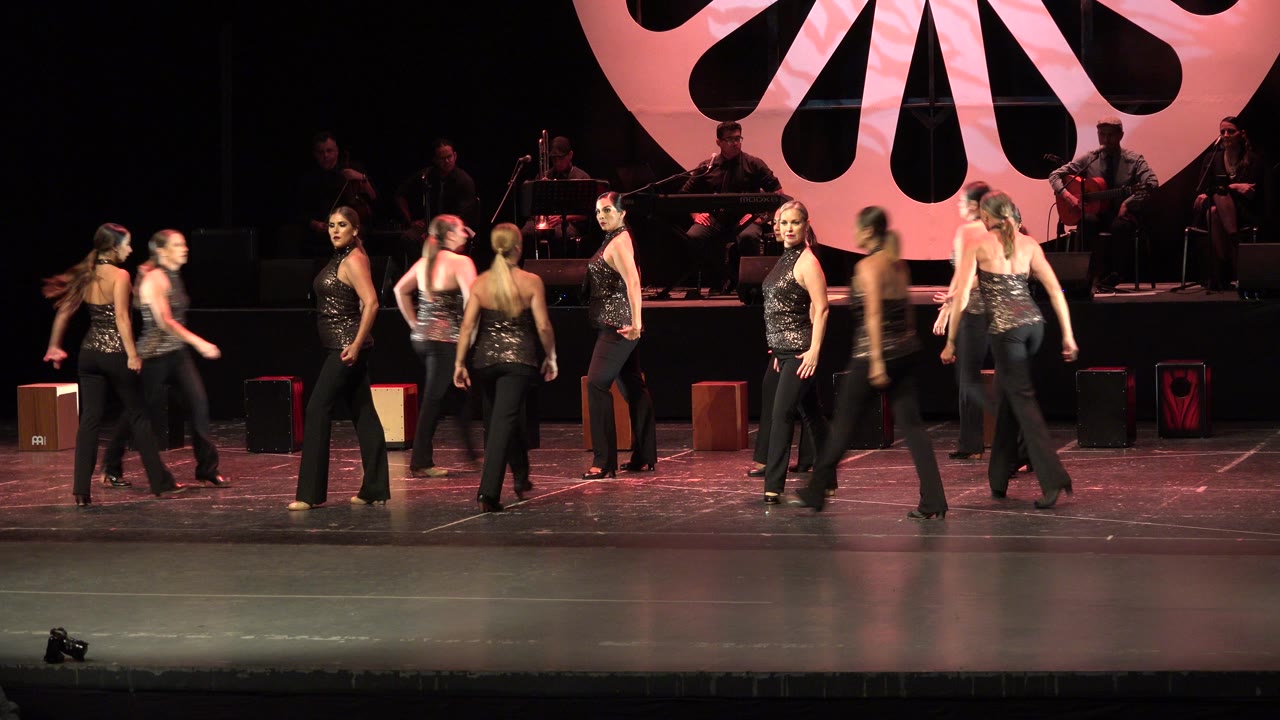 At 0:01, the wide Cabo San Lucas stage view shows several performers walking through positions with seated musicians partly visible in shadow behind them.