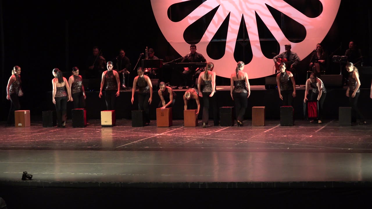 Performers in black stand across a stage with box props and a pink circular backdrop.