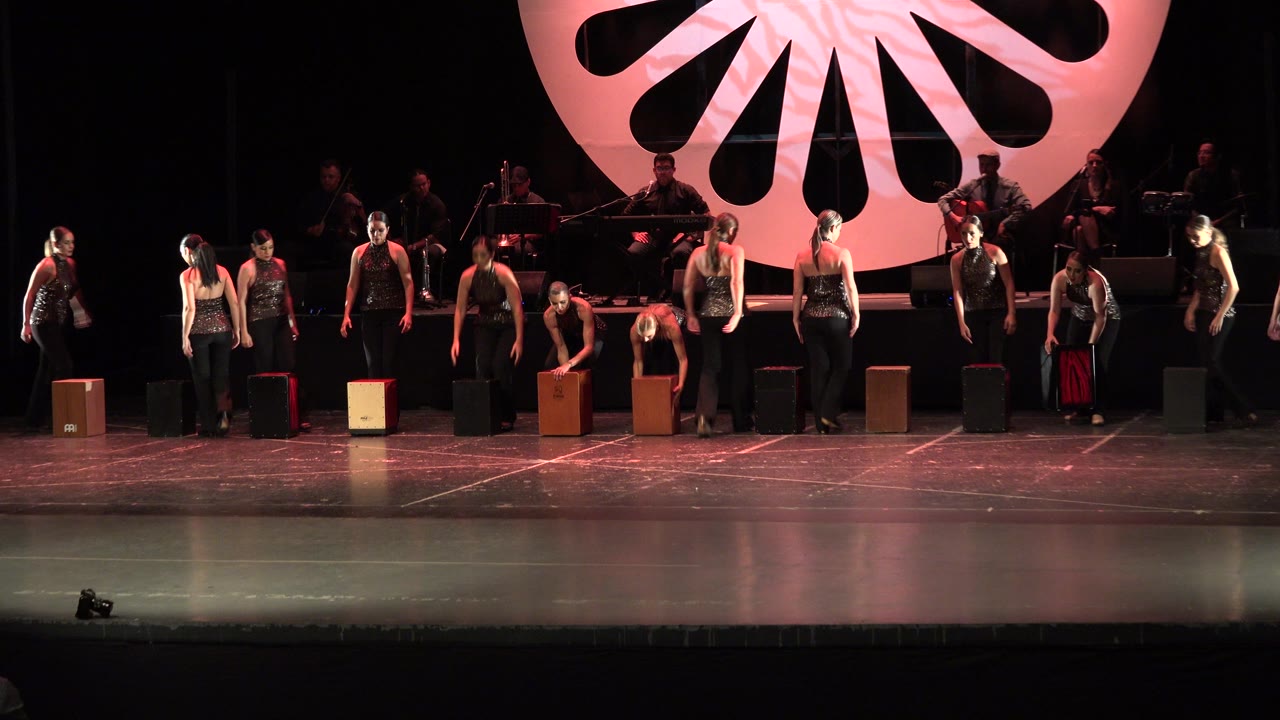 A wider group of performers in black remains arranged across the Cabo San Lucas stage while box props sit in front of several dancers under pink stage light.