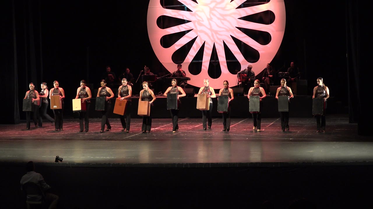The Cabo San Lucas stage performance continues with the row of women spaced across the stage, boxes still held upright under warm pink lighting.