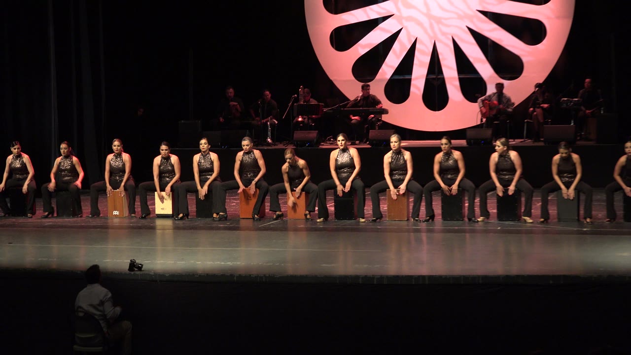 Wide stage view of women seated on wooden box drums with musicians behind them.