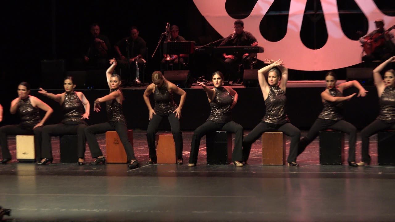 In Cabo San Lucas, performers in black costumes sit in a line on stage while shadowed musicians and a pink circular backdrop fill the rear of the frame.