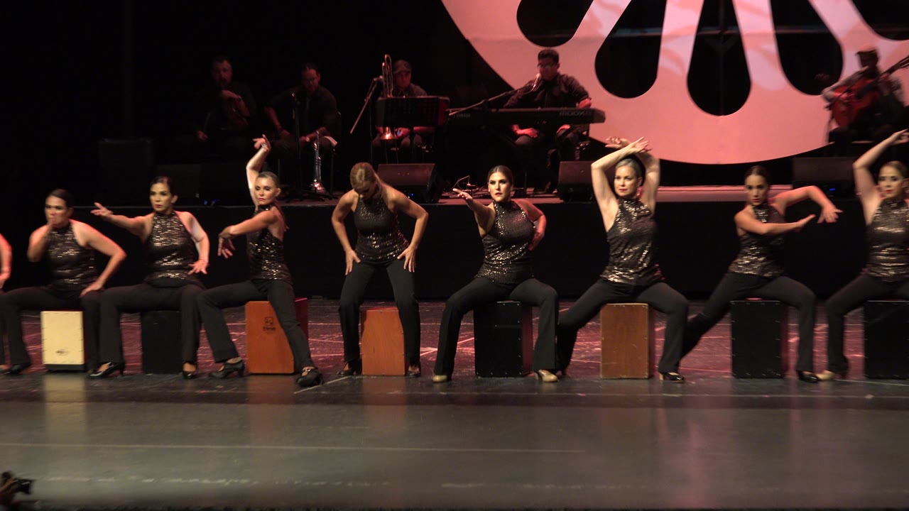 In Cabo San Lucas, the seated performers hold angular arm poses across the stage, with the band partially visible behind them under low warm lighting.