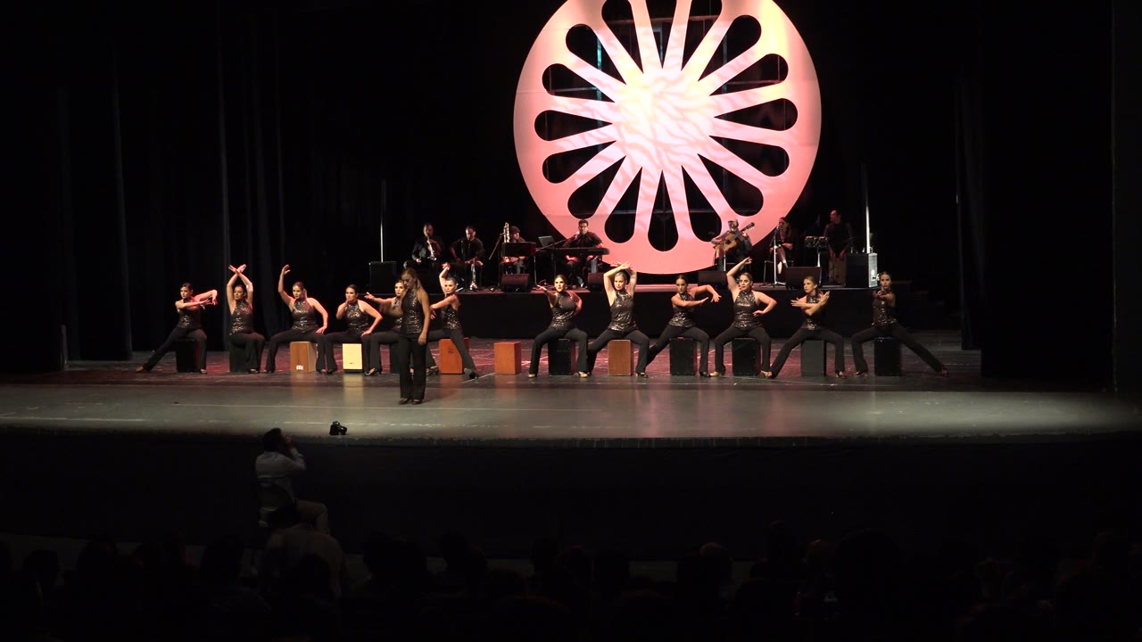 Cabo San Lucas stage performance with a central dancer stepping forward as other dancers pose beside cajon boxes and musicians under the pink set piece.