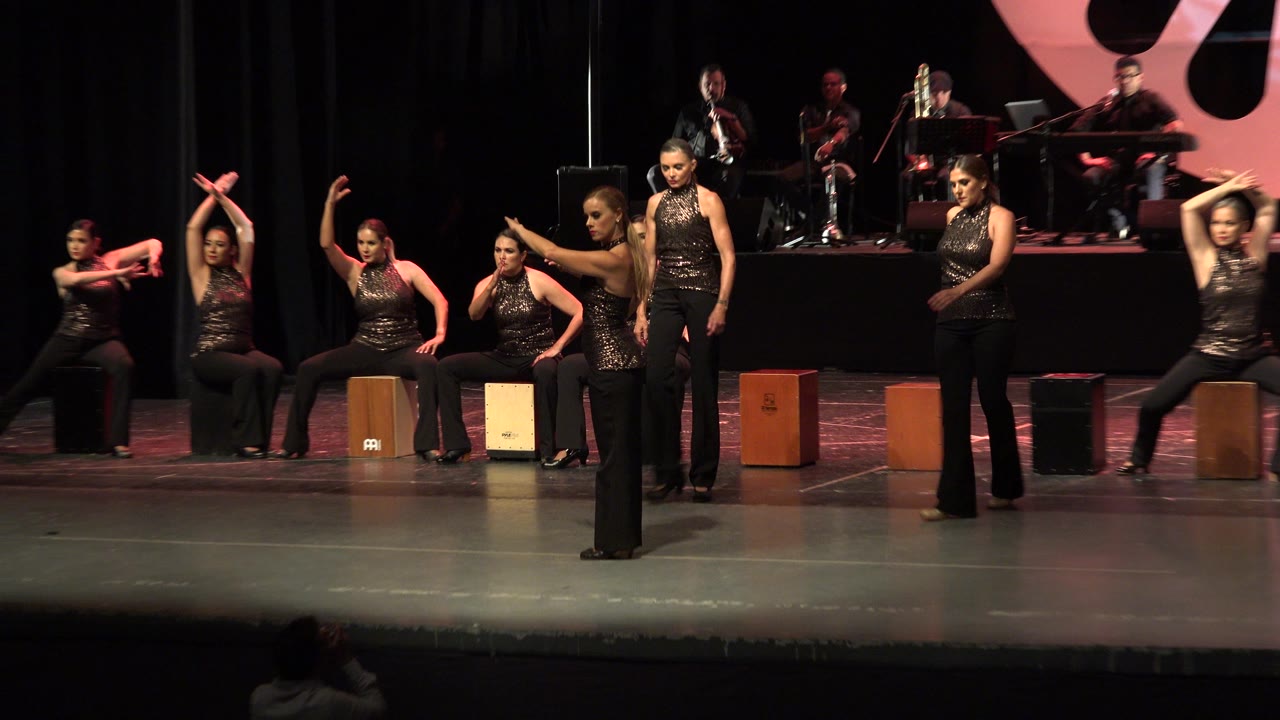 Women performing a dance formation on a theater stage with musicians behind them.