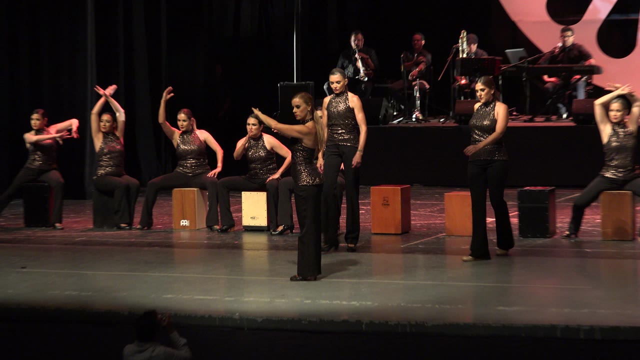 Women on a Cabo San Lucas stage hold arranged dance poses, with several seated near wooden cajon boxes and musicians visible in dim red light behind them.