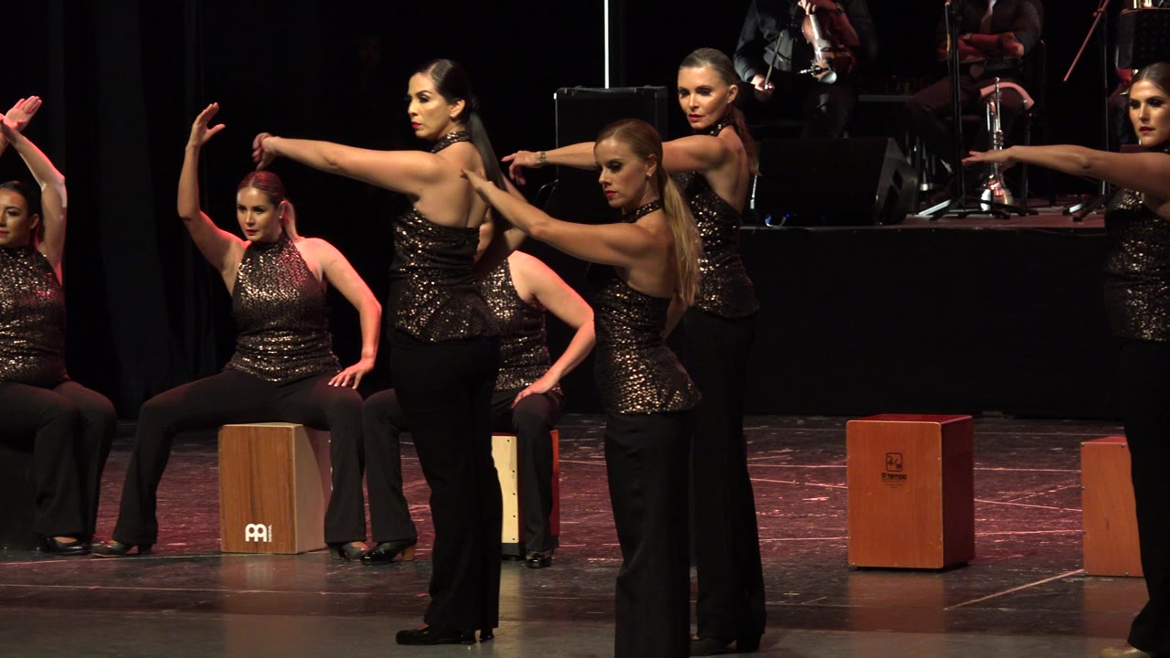 Female dancers hold poses on a dark stage with cajon boxes behind them.