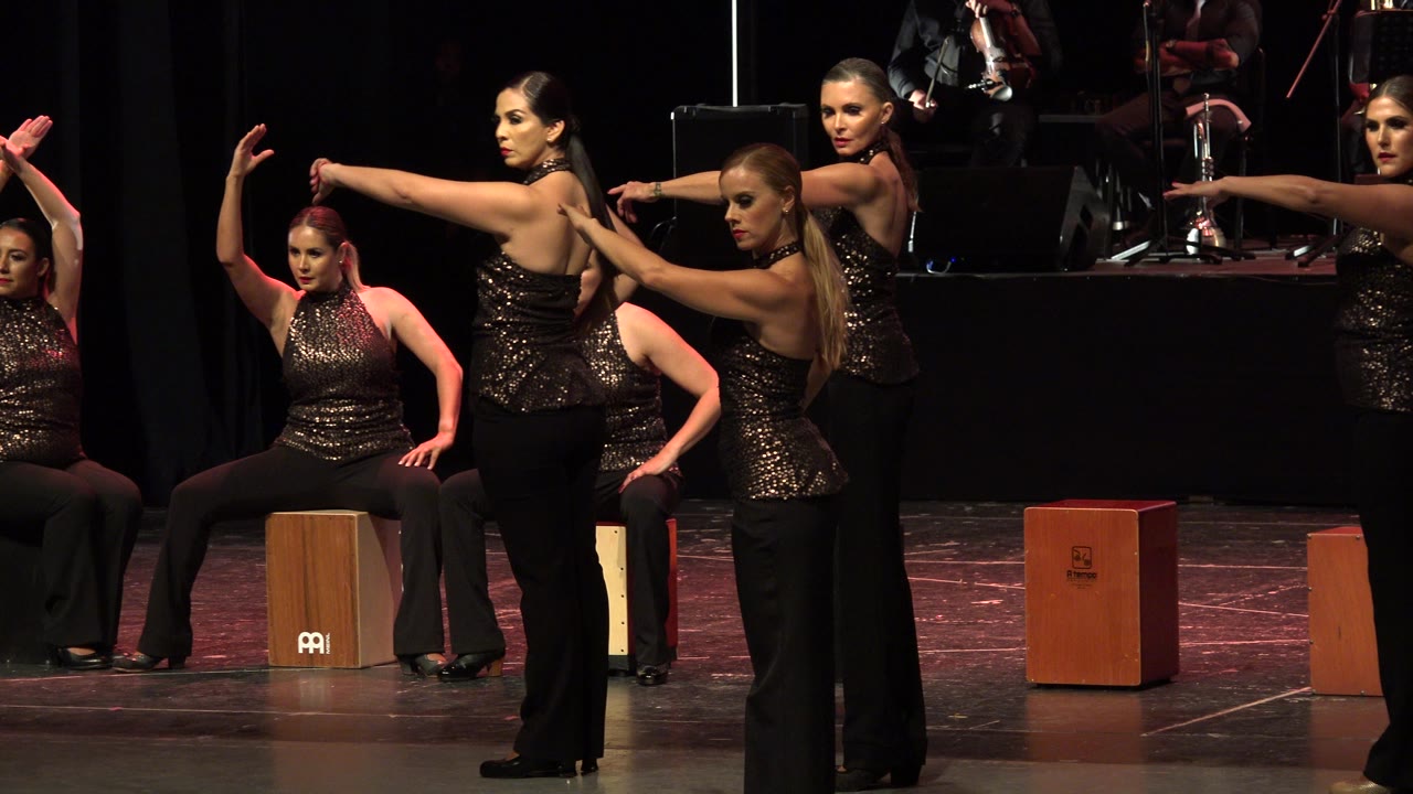 Female performers in black pants and metallic tops extend their arms across the foreground while seated dancers and percussion boxes remain in the background on the Cabo San Lucas stage.
