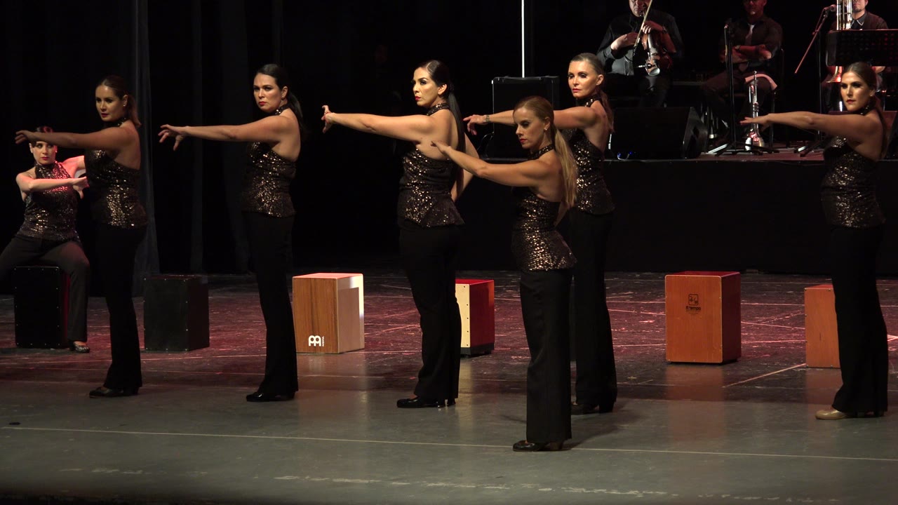 Women in black stage gowns hold extended-arm poses with musicians behind them.